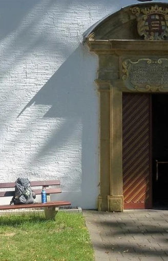 Eingangstor der Kluskapelle St. Lucia umgeben von weißer Wand, Bank und baumbeschattetem Weg.