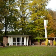 Kaiserbrunnen Brakel Runder Pavillon mit Glasfront im Grünen, umgeben von hohen Bäumen, mit markantem Fahnenmast davor.