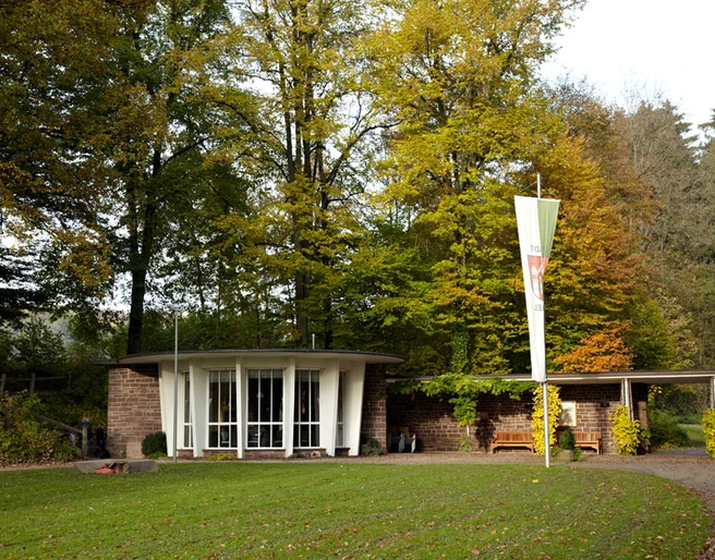 Kaiserbrunnen Brakel Runder Pavillon mit Glasfront im Grünen, umgeben von hohen Bäumen, mit markantem Fahnenmast davor.