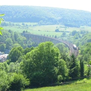 Graues Viadukt schlängelt sich durch grüne bewaldete Hügellandschaft unter blauem Himmel.