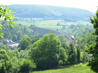 Graues Viadukt schlängelt sich durch grüne bewaldete Hügellandschaft unter blauem Himmel.