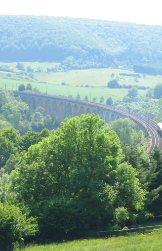 Graues Viadukt schlängelt sich durch grüne bewaldete Hügellandschaft unter blauem Himmel.