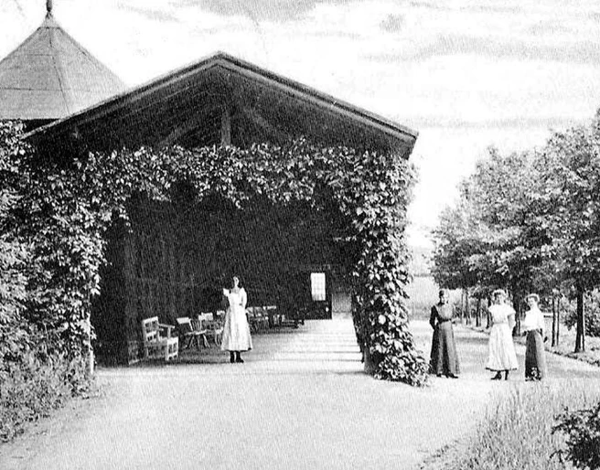 Caspar-Heinrich-Quelle  Historisches Foto eines Pavillons mit Rankpflanzen, umgeben von Frauen in Kleidung des 19. Jahrhunderts.