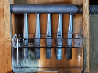 Wasserpfeifen Fünf Metallrohre hängen über einem mit Wasser gefüllten Glasbehälter in einem hölzernen Rahmen.