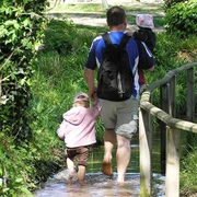 Barfusspark Lienen Ein Mann mit zwei kleinen Kindern läuft barfuß durch einen Wasserweg mit üppigem Grün drumherum.
