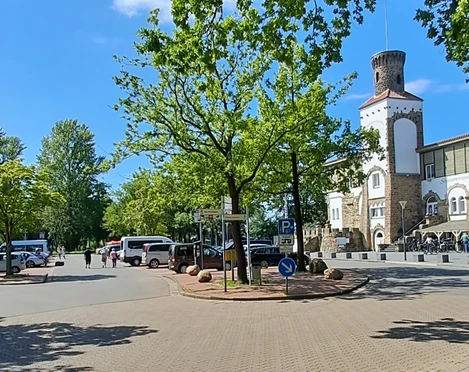 Parkplatz in Strandnähe mit Schatten spendenden Bäumen und einem historischen Turm im Hintergrund.