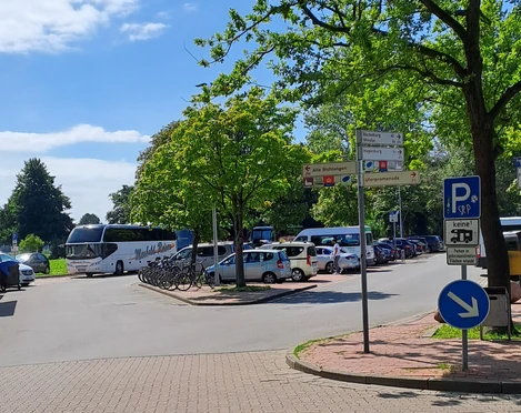 Parkplatz mit gekennzeichneten Flächen für Busse und Fahrräder, umgeben von grünen Bäumen.