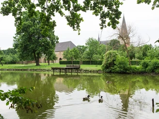 Teich im Garten der Klosteranlage Herzebrock Ruhiger Teich mit Enten im Vordergrund, alte Kirche und gepflegte Gärten im Hintergrund.