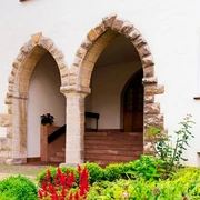 Zukunftswerkstatt Ökumene Warburg-Germete Zwei steinerne Rundbogenportale in einer weißen Fassade, umgeben von gepflegten Blumenbeeten.