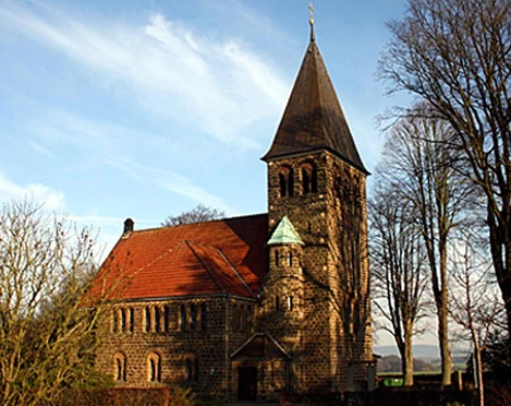 Historische Kirche Hagedorn aus Stein mit rotem Dach, umgeben von Bäumen unter blauem Himmel.