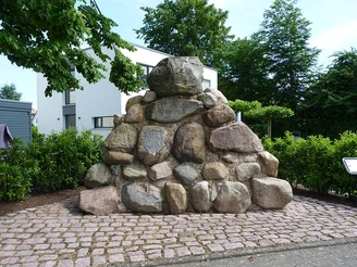 Steinhaufen-Denkmal aus glatten, großen Felsen, umgeben von grünen Hecken und Bäumen.