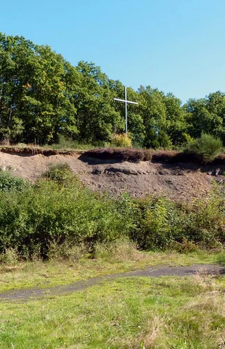 Spatzenberg Holzweg durch eine grasbewachsene Lichtung, umgeben von grünem Wald und einem Holzkreuz.