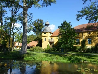 Schloss Gut Böckel mit gelben Wänden und einem Turm inmitten grüner Bäume reflektiert im Wasser.