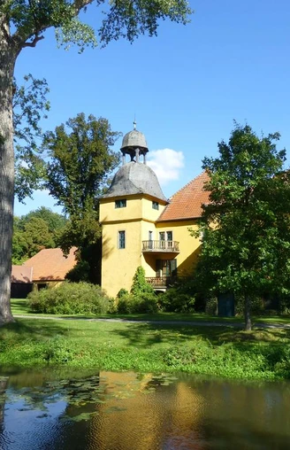 Schloss Gut Böckel mit gelben Wänden und einem Turm inmitten grüner Bäume reflektiert im Wasser.