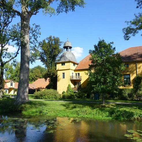 Gut Böckel Schloss Gut Böckel mit gelben Wänden und einem Turm inmitten grüner Bäume reflektiert im Wasser.
