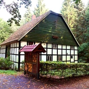 Katzenholz - Hühnerhaus Fachwerkhaus im Grünen, umgeben von Bäumen, mit Schindeldach und dekorativem Holzportal im Vordergrund.