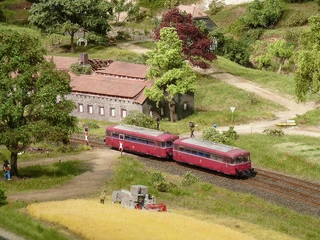 Modellbundesbahn Schienenbus im Weserbergland Ein roter Schienenbus passiert eine ländliche Modelllandschaft mit einem Fachwerkgebäude und Feldern.