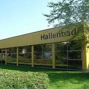 Städtisches Hallenbad Gelbes Gebäude eines städtischen Hallenbades mit großen Fenstern, umgeben von grüner Landschaft.