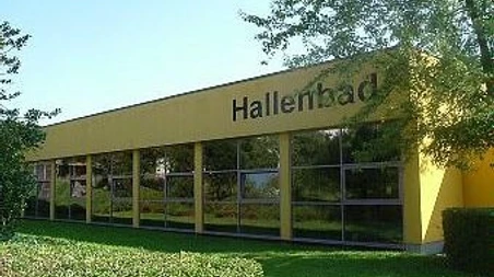 Städtisches Hallenbad Gelbes Gebäude eines städtischen Hallenbades mit großen Fenstern, umgeben von grüner Landschaft.