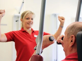 Training Frau in rotem Polo-Hemd leitet älteren Mann beim Krafttraining an einer Fitnessmaschine an.