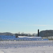 Blick auf die ehemalige Knochenleimfabrik Schneebedeckte Felder mit bäuerlichen Gebäuden und einem Schornstein. Winterlandschaft unter blauem Himmel.