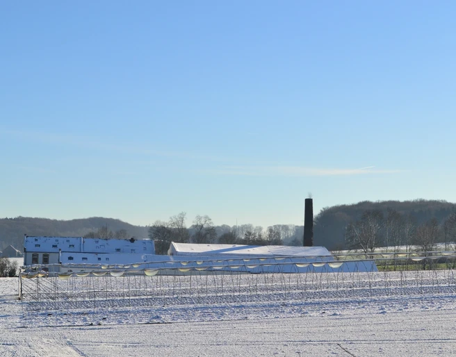 Blick auf die ehemalige Knochenleimfabrik Schneebedeckte Felder mit bäuerlichen Gebäuden und einem Schornstein. Winterlandschaft unter blauem Himmel.