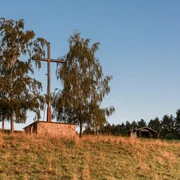 Ein großes Holzkreuz neben zwei Bäumen auf einem grasbewachsenen Hügel vor klarem, blauem Himmel.