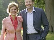 Ein lächelndes Paar spaziert in formeller Kleidung durch einen grünen Park bei Sonnenlicht.