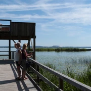 Aussichtsplattform Moorhütte Ein Paar genießt die Aussicht auf einem hölzernen Steg mit Plattform, umgeben von einem See und Segelbooten.
