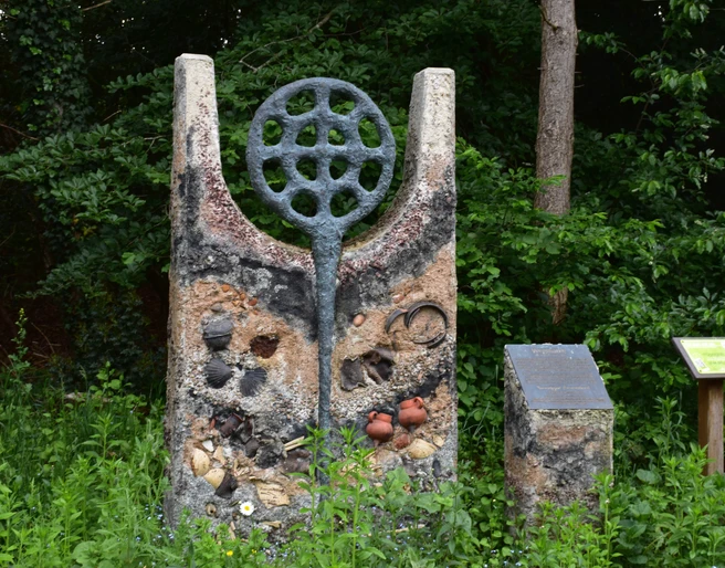 Hügelgrab Das Bild zeigt eine steinerne Skulptur mit kreisrunden und parallel angeordneten Elementen sowie einem angrenzenden Infotafel im Grünen.