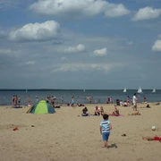 Ein sonniger Strand voller Menschen mit bunten Badeanzügen und bunten Schirmen am Steinhuder Meer. Segelboote gleiten ruhig auf dem Wasser.