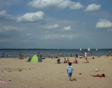 Badeinsel Steinhude Ein sonniger Strand voller Menschen mit bunten Badeanzügen und bunten Schirmen am Steinhuder Meer. Segelboote gleiten ruhig auf dem Wasser.
