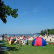 Strandleben auf der Badeinsel Mehrere Personen genießen sommerliche Aktivitäten auf einer grünen Wiese nahe einem sandigen Strand.