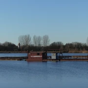 Baggersee zw. Anreppen und Delbrück Blauer See mit Saugbagger im Vordergrund, Ufer mit Bäumen und einem Kirchturm im Hintergrund.
