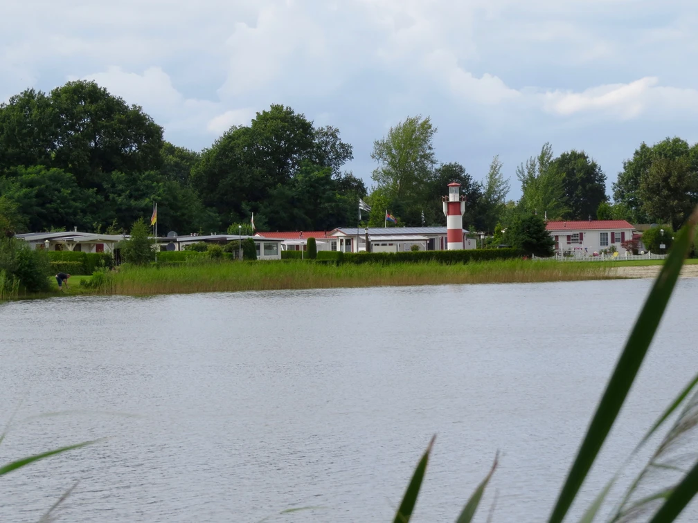 Ein idyllischer Campingplatz am Ufer eines Sees, geprägt von grüner Vegetation und einem markanten rot-weißen Leuchtturm im Hintergrund.