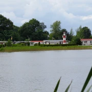 Ein idyllischer Campingplatz am Ufer eines Sees, geprägt von grüner Vegetation und einem markanten rot-weißen Leuchtturm im Hintergrund.