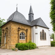Kleine historische Kapelle mit schiefergedecktem Dach, flankiert von Bäumen und einem klaren Himmel.