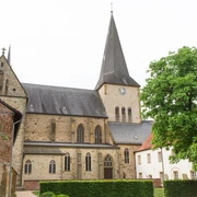 Historische Kirche mit hohem Turm und Uhr, umgeben von alten Gebäuden und grünem Baum.