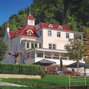 Villa Thusnelda mit dem Café Bistro Daheim Ein weißes Gebäude mit rotem Dach und Uhrturm, umgeben von Bäumen, beherbergt das Café Richter; davor stehen Tische mit Sonnenschirmen.