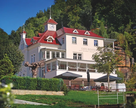 Villa Thusnelda mit dem Café Bistro Daheim Ein weißes Gebäude mit rotem Dach und Uhrturm, umgeben von Bäumen, beherbergt das Café Richter; davor stehen Tische mit Sonnenschirmen.