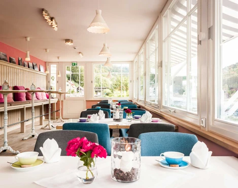 Café Bistro Daheim Innenraum Heller, gemütlicher Café-Raum mit großen Fenstern, blauen Stühlen, rosa Kissen und dekorativen Blumen auf den Tischen.