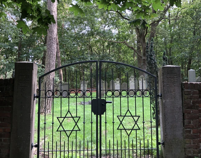 Jüdischer Friedhof  Historischer jüdischer Friedhof mit schwarzem Eisentor, verzierten Davidsternen und grünem Baumhintergrund.