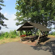 Blick auf den Picknickplatz in der Schutzhütte am Hagenburger Kanal Bastion 2 Picknickplatz mit Holztisch und Bänken unter einem überdachten Unterstand, umgeben von grüner Natur.