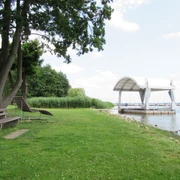 Ein idyllischer Picknickplatz am Ufer mit grüner Wiese, Bäumen und angrenzender Seebrücke.
