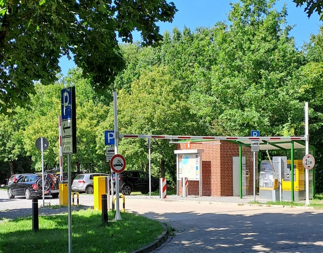 Kassenautomat am Parkplatz Bruchdamm, umgeben von Bäumen und parkenden Autos bei sonnigem Wetter.