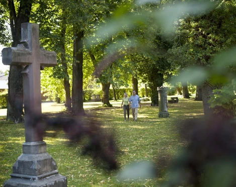 hx-stadtentdecken-andreashub-7 Pärchen auf dem lichtdurchfluteten und mit Bäumen bewachsenem Friedhofswall mit großem Sandsteinkreuz