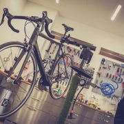 Fahrradwerkstatt-Platzhalter In einer Fahrradwerkstatt hängt ein schwarzes Rennrad auf einem Ständer, umgeben von Werkzeugen an einer Werkbank.