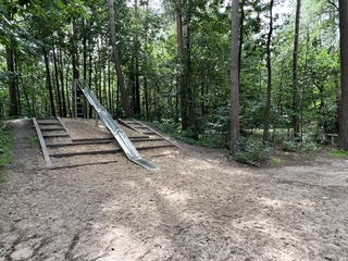 Waldspielplatz Kirchlinteln Eine Rutsche im Wald, umgeben von hohen Bäumen, steht auf einem sandigen Spielplatzboden.