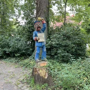 Röbenmarktmann Holzskulptur eines lächelnden Marktschreiers mit blauer Kleidung, auf einem Baumstumpf im Grünen.