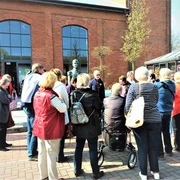 g-stef-hrung-zeitreise-augustfehn-apen Eine Gruppe von Menschen steht vor einem Backsteingebäude und hört einem Redner zu.